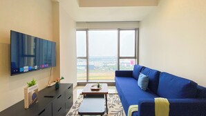 A modern living room with a large TV, a blue sofa, a small coffee table, and a view of the city through large windows.