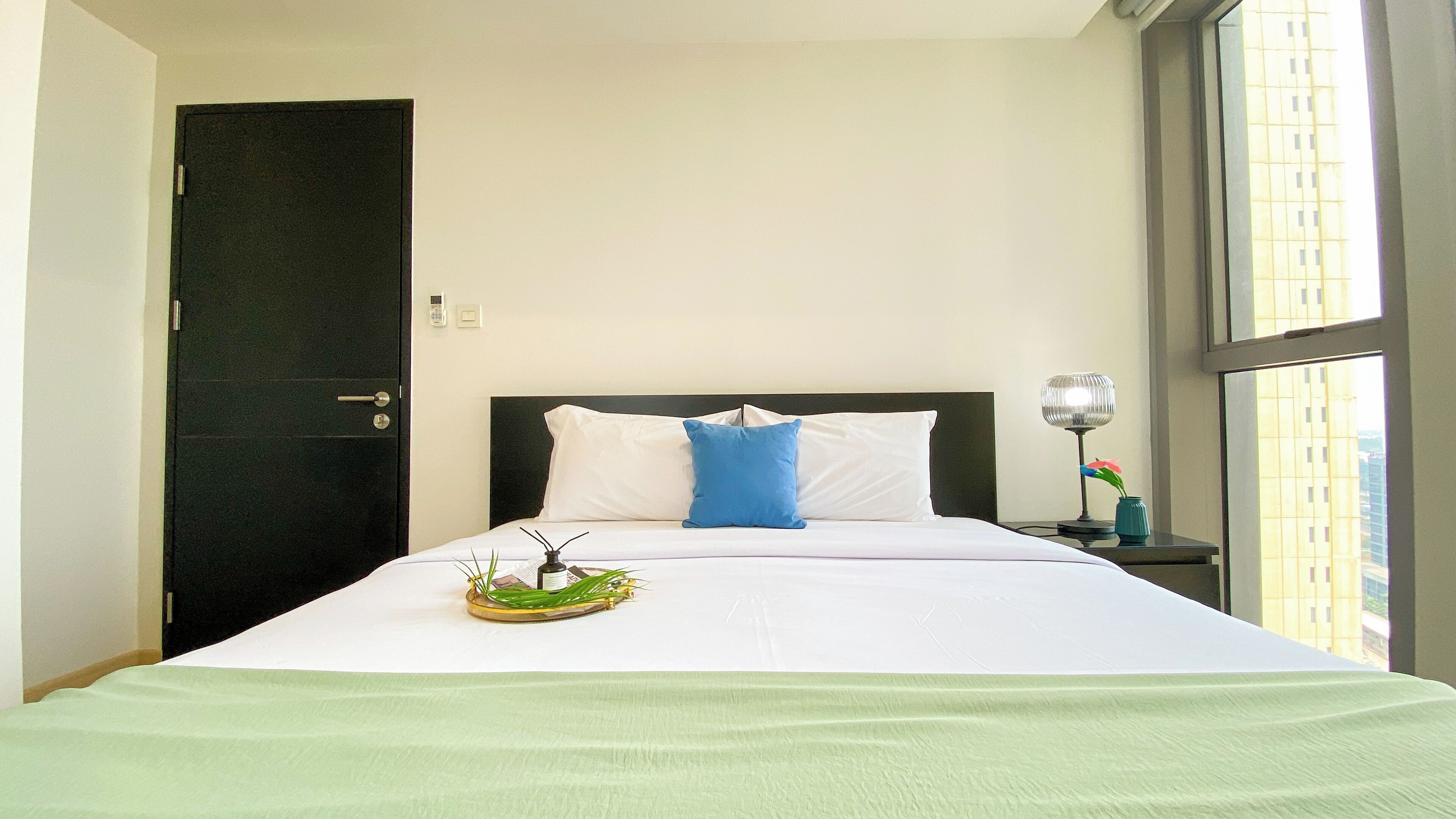 Een net opgemaakt bed met witte en blauwe kussens, een nachtkastje met een lamp, en een raam met uitzicht op gebouwen.