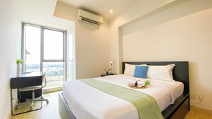 A hotel room with a large bed, a desk with a chair, a window with a view, and an air conditioning unit.