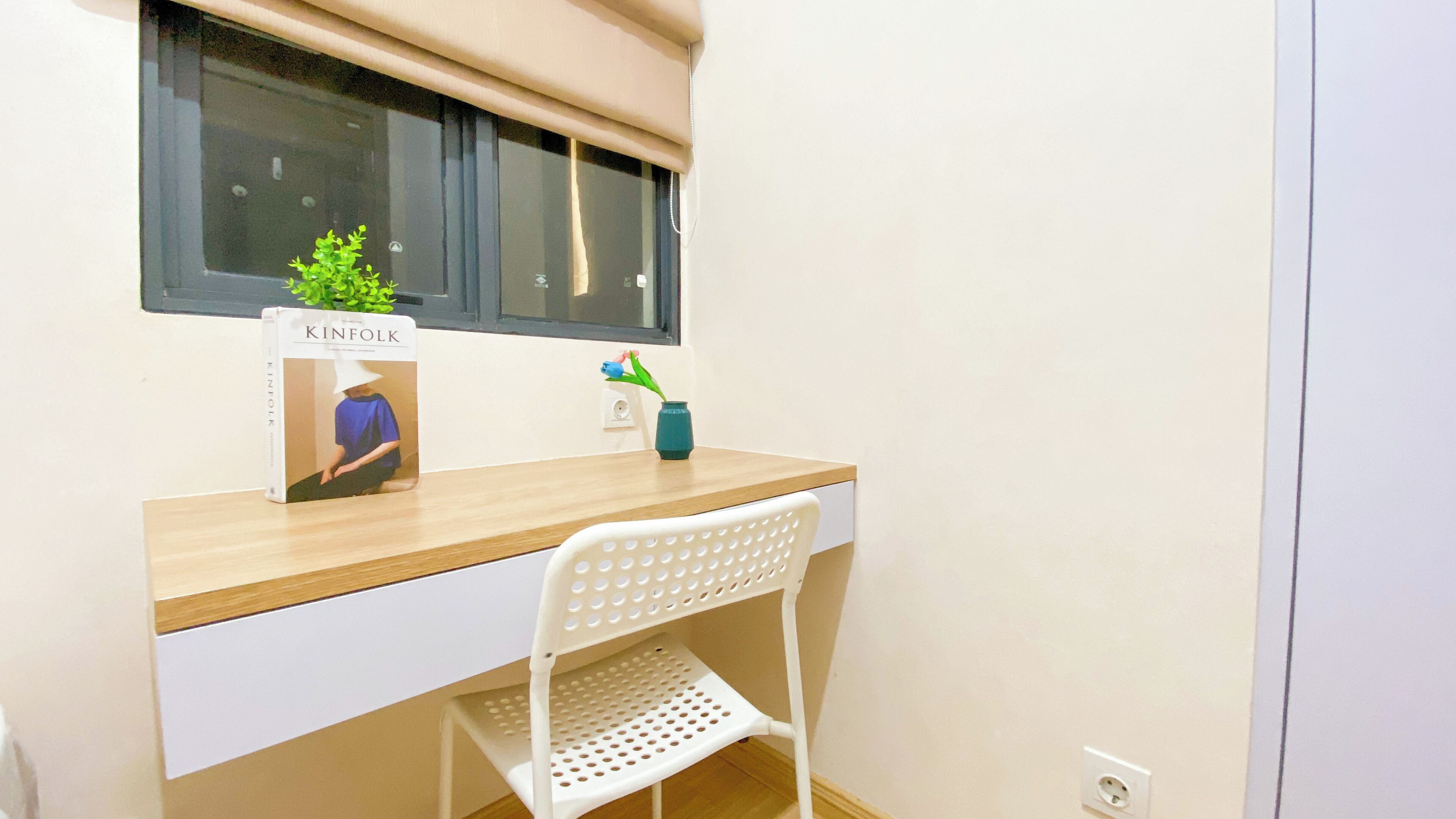 A small, tidy room with a wooden desk, a white chair, a window with a blind, and a small plant on the desk.