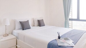 A neatly made bed with white bedding and two pillows, a bedside table with a lamp, and a window with light blue curtains. - Best Place And Homey 2Br Sky House Bsd Apartment Near Aeon (Pagedangan)