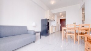 A modern living room with a grey sofa, a black refrigerator, a wooden dining table with chairs, and a small side table with a lamp. - Best Place And Homey 2Br Sky House Bsd Apartment Near Aeon (Pagedangan)