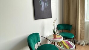 Une pièce avec deux fauteuils verts, une petite table ronde surmontée d’un coussin rayé aux couleurs vives, et une orchidée en pot.