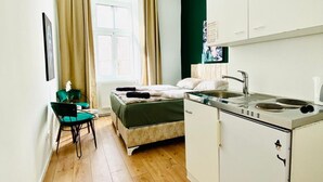 Une pièce de taille modeste comprenant un lit, une petite kitchenette, un coin repas avec une chaise et une fenêtre avec des rideaux.