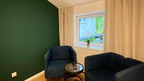 Ett rum med två mörkfärgade fåtöljer, ett litet runt bord, ett fönster med gardiner och utsikt över grönområden utanför.
