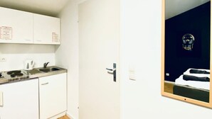 Ett litet pentry med en diskho, en spis och skåp, en dörr med ett handtag och en spegel som reflekterar en säng och en hjälm.