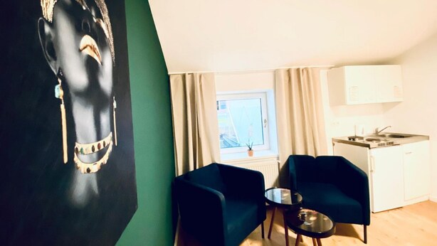 Ett modernt rum med en svart väggdekoration, två blå fåtöljer, ett litet runt bord, ett fönster med gardiner, en liten köksdel och ett element.