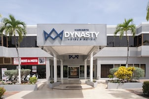 The exterior of the Margaritah Dynasty Hotel & Suites with a modern design, featuring a prominent logo, a covered entrance, and a 'Rent-A-Moto' service sign.