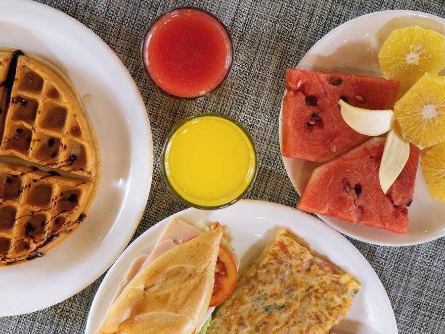 A breakfast plate with waffles, a glass of red juice, a glass of orange juice, watermelon slices, and a sandwich.