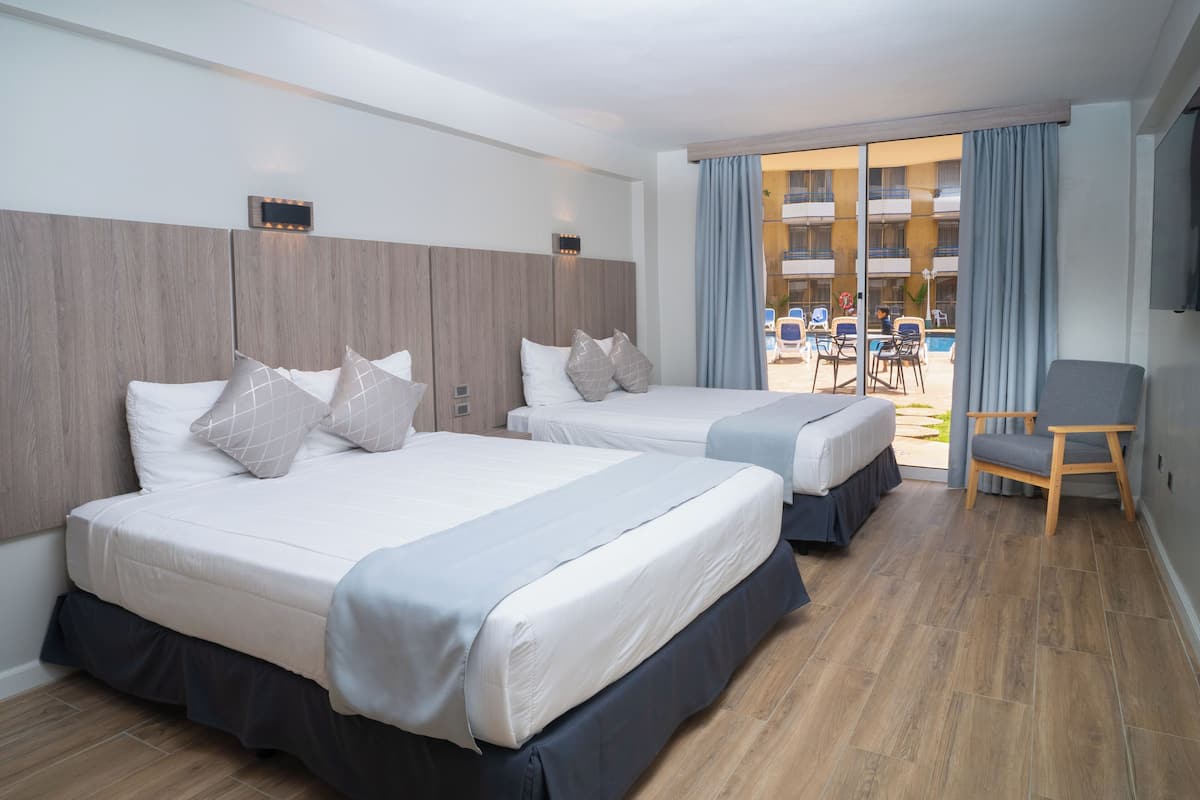 A hotel room with two beds, a wooden floor, a balcony with a pool, and a chair.