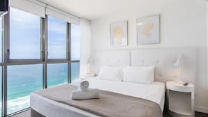 Une chambre d’hôtel moderne dotée d’une grande fenêtre donnant sur la mer, d’un lit soigneusement fait et d’une petite table de chevet avec une lampe.