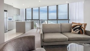 Un salon moderne avec un canapé et une table basse en verre, une cuisine équipée d’un four à micro-ondes et de grandes fenêtres offrant une vue sur la ville.