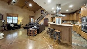 A spacious living area with a large sofa, a coffee table, and a fireplace. There is a kitchen area with wooden cabinets and a stainless steel range hood. A staircase leads to an upper level. - Peaceful Texas Ranch Retreat on the Lake (Midlothian)