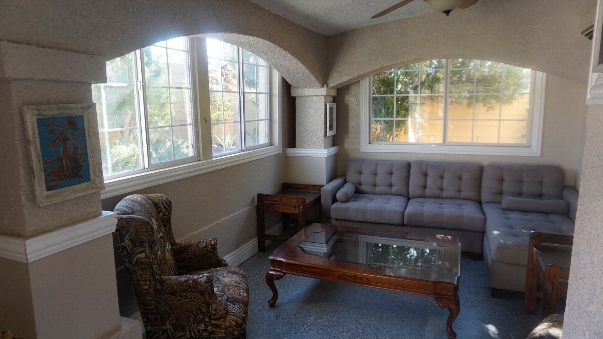 A living room with a sofa, armchair, and coffee table. There are two windows with views of greenery outside.