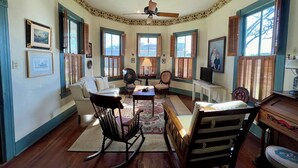 A cozy living room with a wooden floor, a ceiling fan, and a large window with shutters. There is a sofa, a rocking chair, and a small table with a lamp.