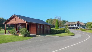 A wooden cabin with a porch and flower pots, a curved driveway, and a large house in the background.