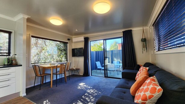 Un espacio habitable compacto con comedor, un sofá con un cojín estampado y una puerta corrediza de vidrio que da a un patio exterior.