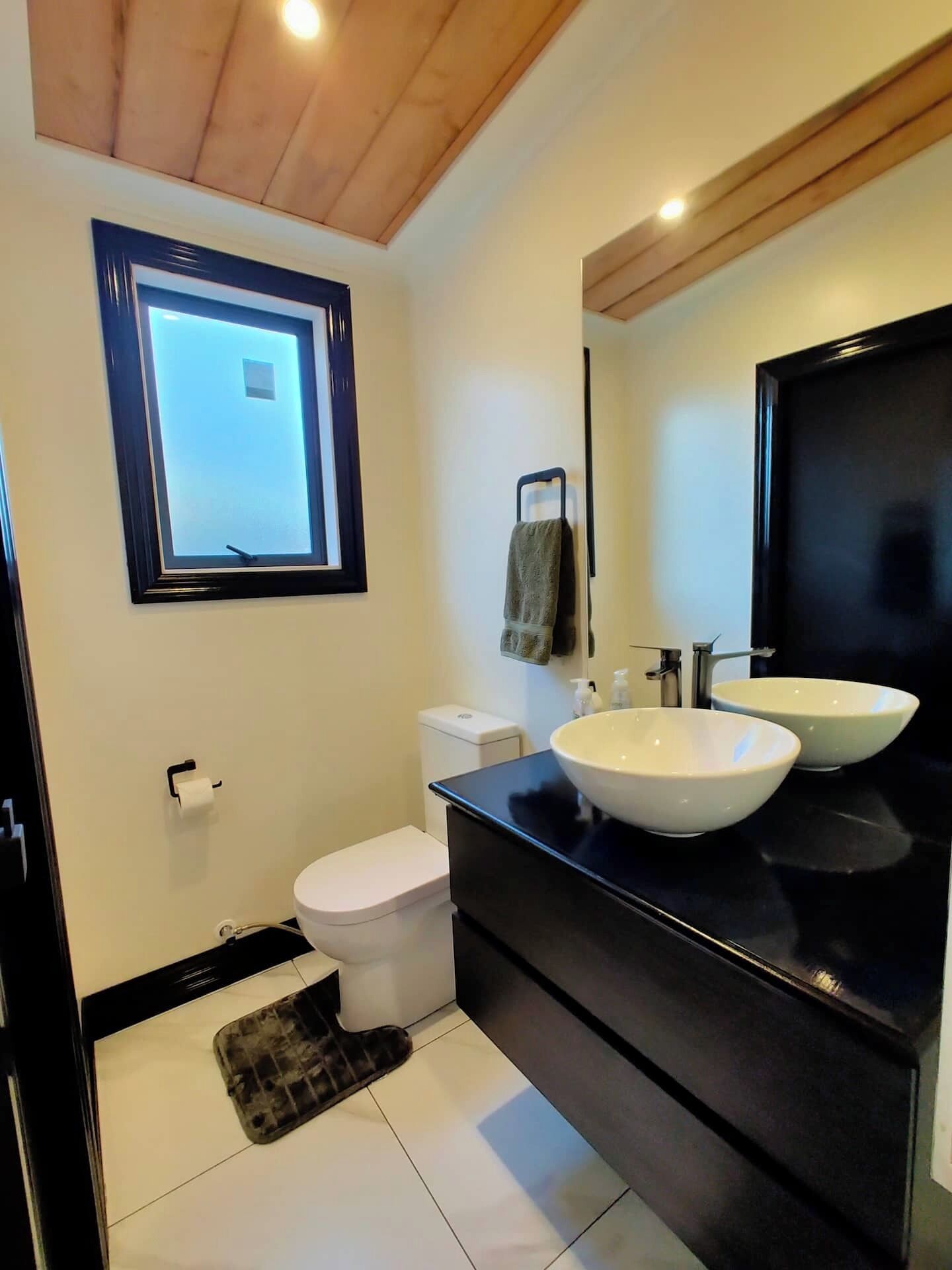 A modern bathroom with a black vanity, two sinks, a toilet, and a window.