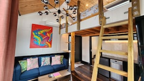 Ein modernes Loft-Zimmer mit Schlafgalerie, einem blauen Sofa, einem farbenfrohen abstrakten Gemälde und einer Holzbalkendecke mit einem einzigartigen Kronleuchter.