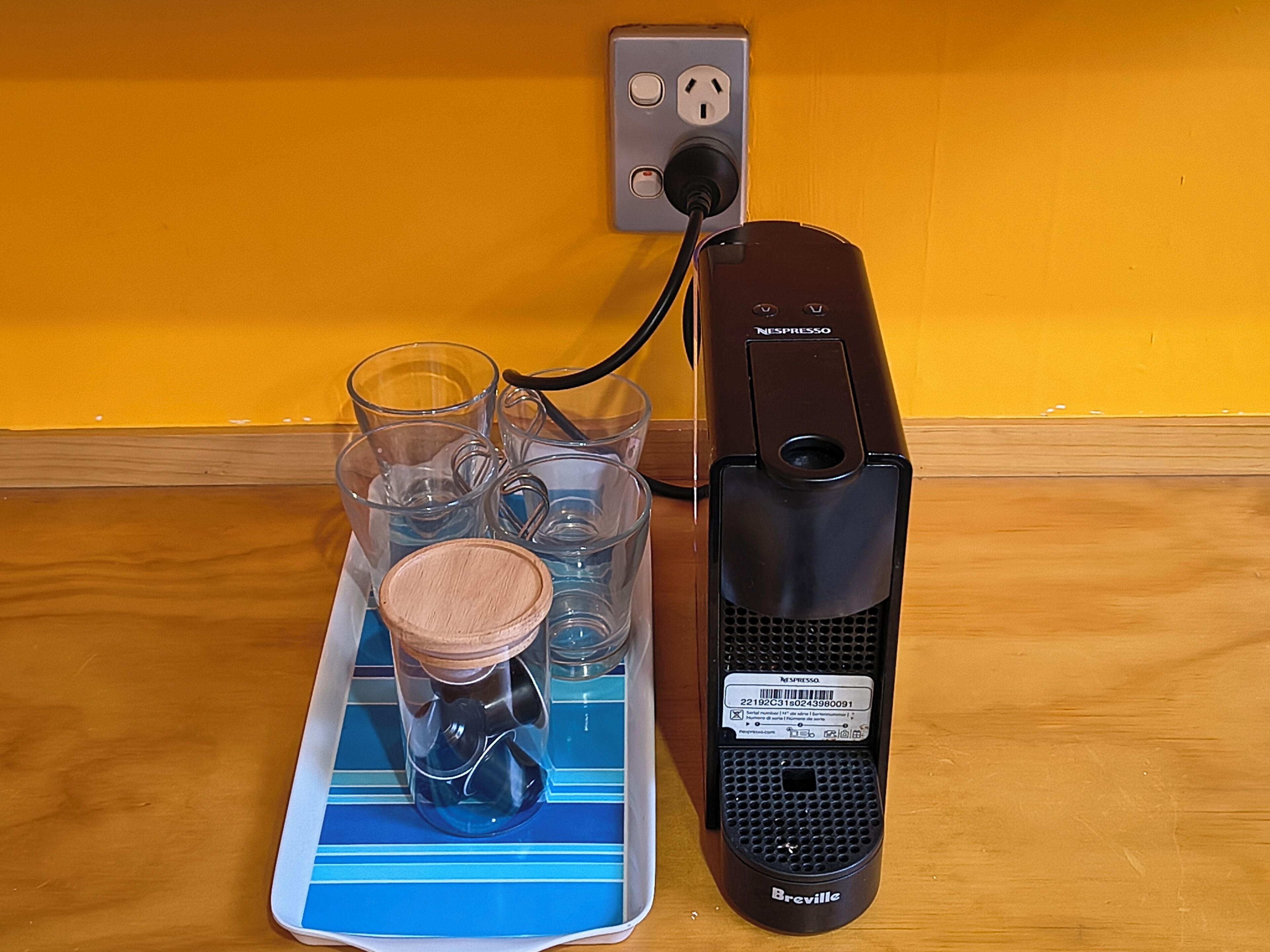 Uma cafeteira Breville conectada a uma tomada, um tabuleiro azul com copos de vidro transparente e uma tampa de madeira, e uma parede amarela.