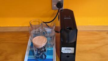 Uma cafeteira Breville conectada a uma tomada, um tabuleiro azul com copos de vidro transparente e uma tampa de madeira, e uma parede amarela.