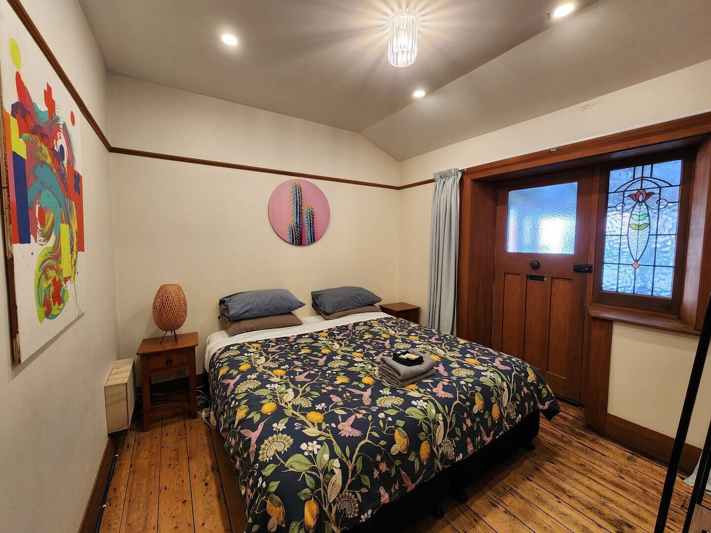A bedroom with a wooden floor, a bed with a floral bedspread, a round wall decoration, a wooden door with a stained glass window, and a small round table with a lamp.
