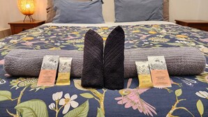 Una cama con colcha estampada, dos almohadas y un par de toallas dobladas. Hay tres botellas de jabón y champú sobre la cama.