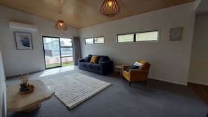 A spacious living room with a grey carpet, a blue sofa, a yellow armchair, and a white rug. There are two pendant lights, a small table with a plant, and a window with a view of the outside. - Sleek Suite by the Sand (2-bedroom) (Dunedin)