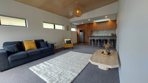 A modern living room with a sofa, coffee table, and a rug. There is a kitchen area with stools and a bar counter. - Sleek Suite by the Sand (2-bedroom) (Dunedin)