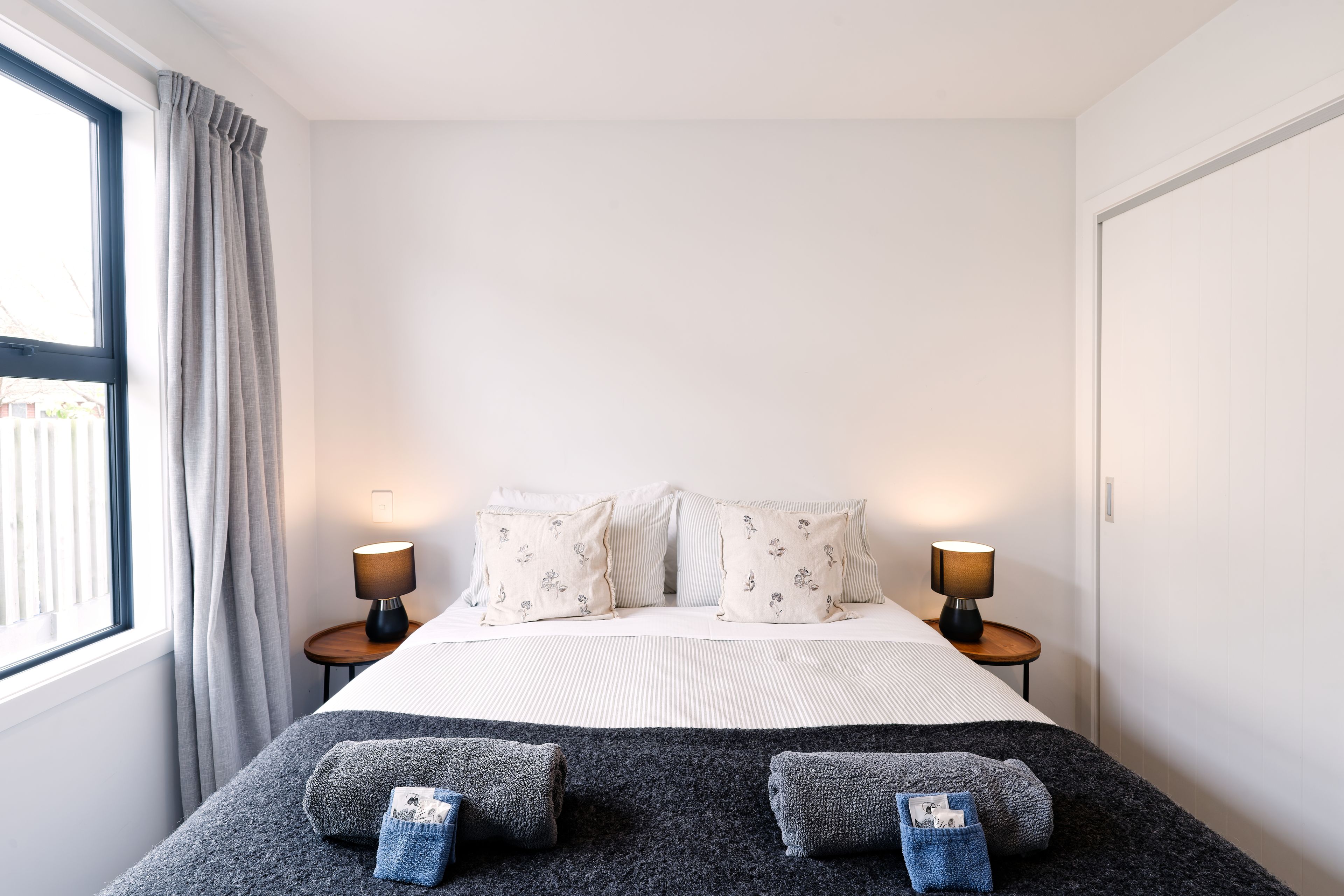 A bedroom with a bed, two bedside lamps, a window with curtains, and a view of the outside.