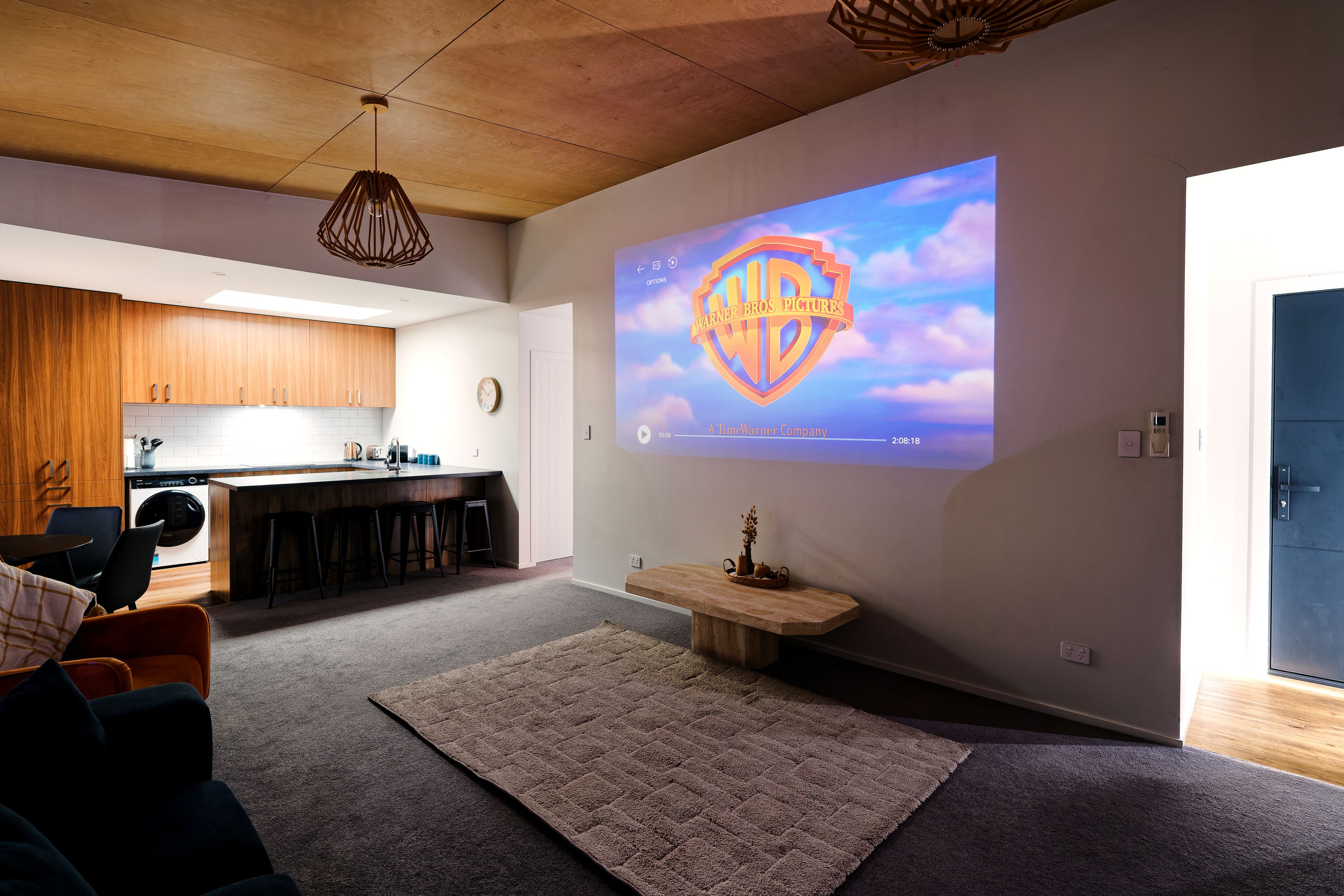 Una sala de estar moderna con una pantalla grande que muestra el logo de una película, una cocina al fondo y un rincón de descanso acogedor.