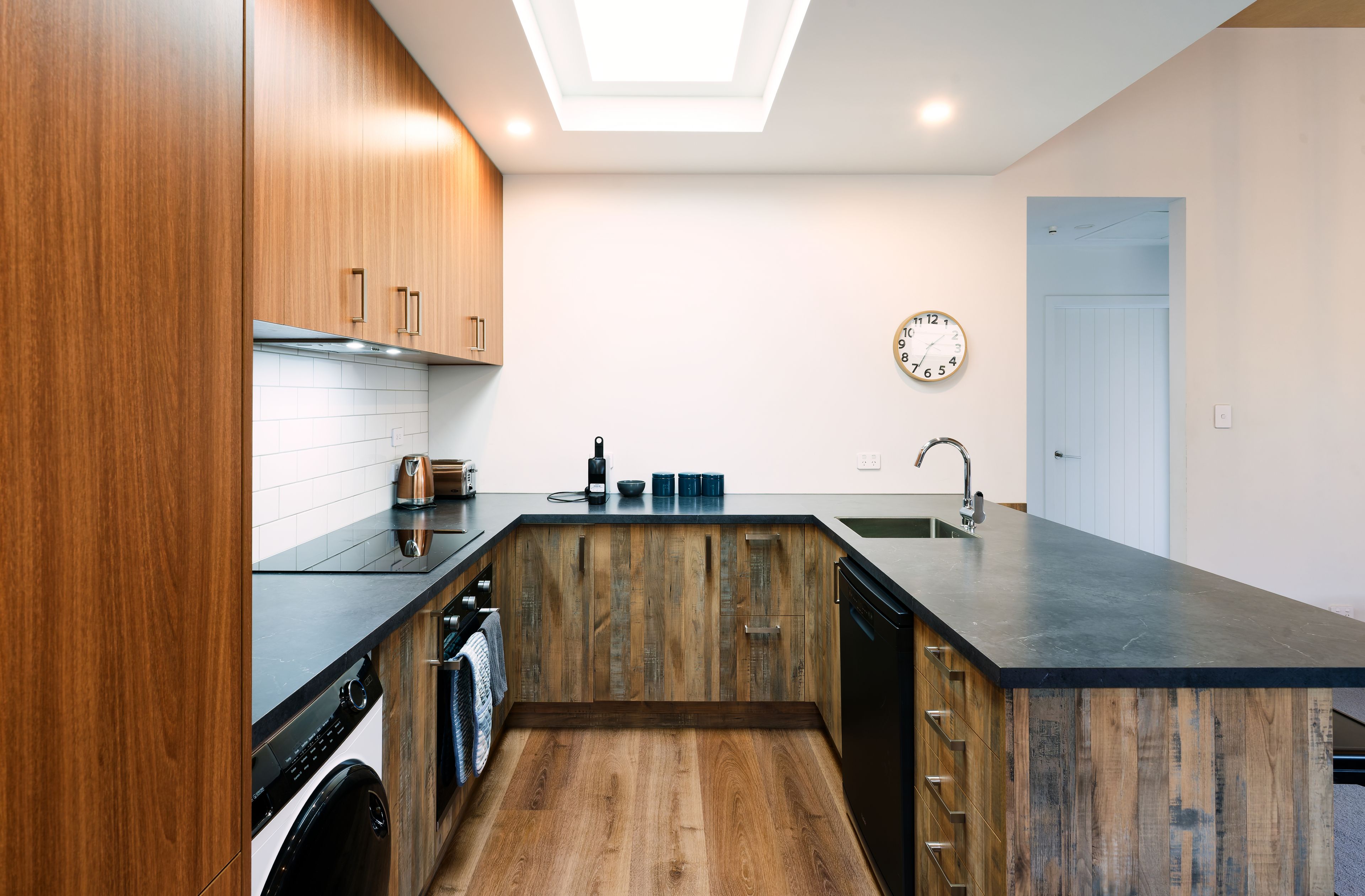 A modern kitchen with wooden cabinetry, a black countertop, a built-in dishwasher, and a skylight above.