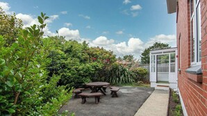 A patio with picnic tables, surrounded by lush greenery and a brick building with a glass door. - Roslyn Retreat: Garden 2-Bed Suite + Kitchenette (Dunedin)