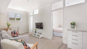 A modern, well-lit room with a sofa, a TV, a small table with a vase, and a bed in the background. - Roslyn Retreat: Garden 2-Bed Suite + Kitchenette (Dunedin)