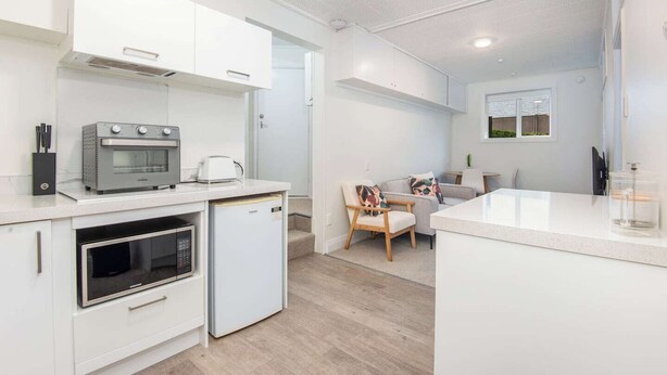 A modern kitchen with white cabinetry, a built-in oven, a microwave, and a small refrigerator. There is a dining area with a table and chairs, and a window with a view. - Roslyn Retreat: Garden 2-Bed Suite + Kitchenette (Dunedin)