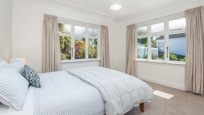 A bedroom with a bed, two windows with curtains, and a view of the garden and house outside.