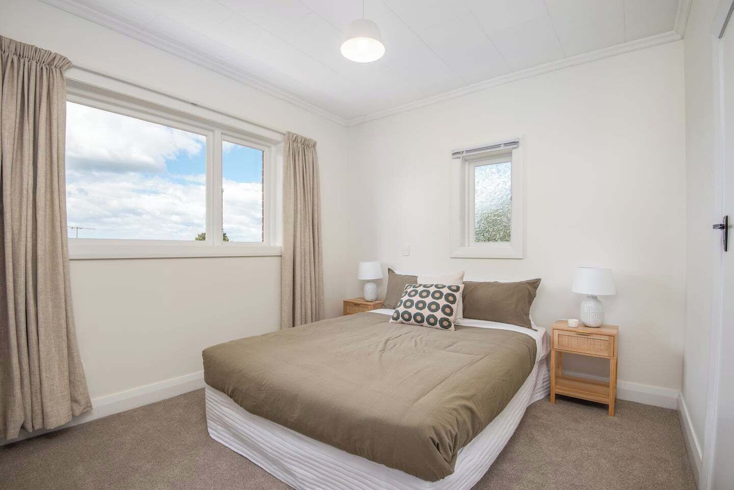 A bedroom with a bed, two bedside tables with lamps, a window with curtains, and a view of the sky.