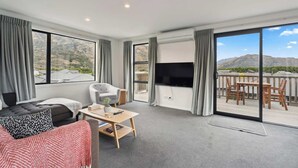 A modern living room with a sofa, armchair, and a table. A TV is mounted on the wall, and there is a balcony with a table and chairs.