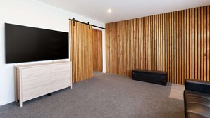 Un soggiorno moderno con un ampio televisore a schermo piatto, un pannello parete in listelli di legno, un tappeto grigio, un divano nero e un mobiletto in legno.