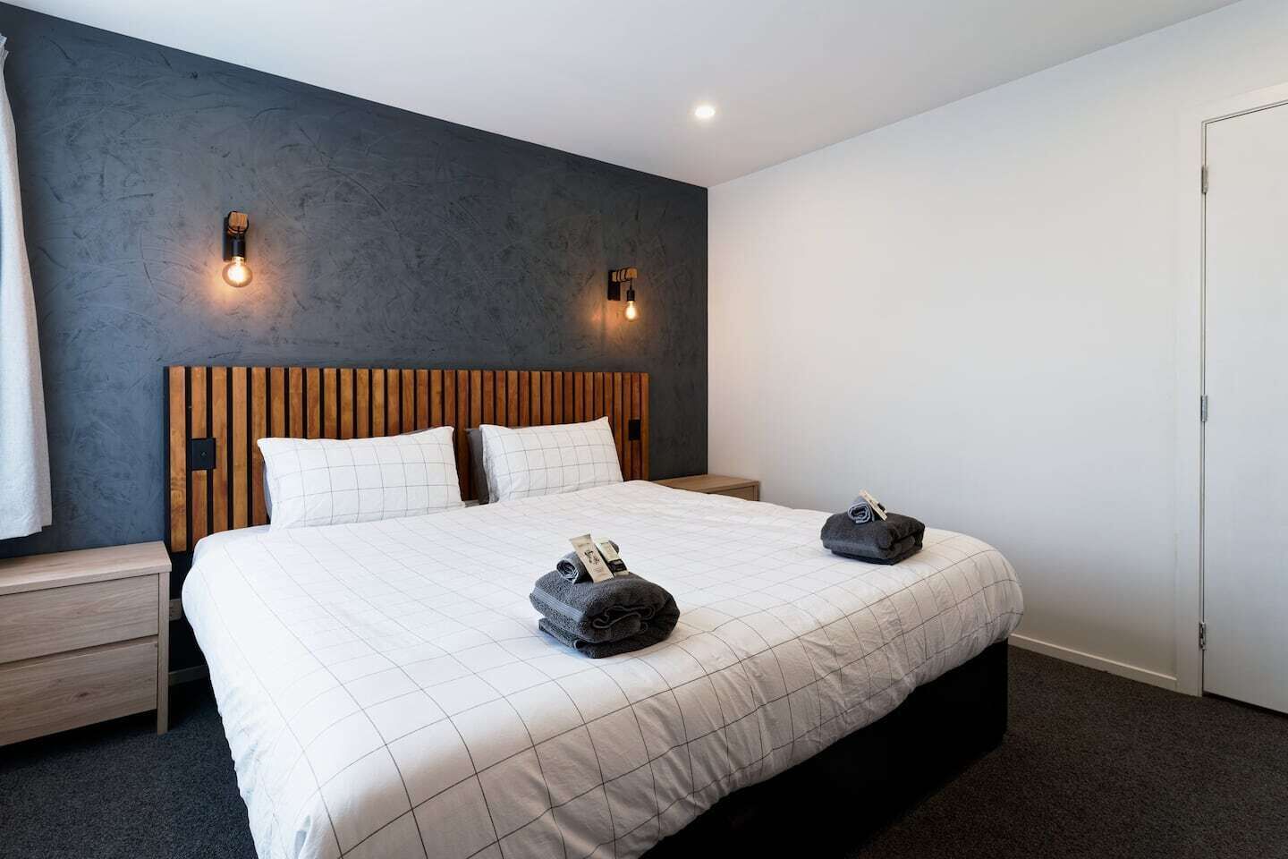 A modern hotel room with a large bed, wooden headboard, two bedside tables, and a dark accent wall with wall sconces.