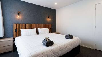 A modern hotel room with a large bed, wooden headboard, two bedside tables, and a dark accent wall with wall sconces.