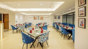 A spacious dining area with multiple tables and blue chairs, featuring a clean, modern design with recessed lighting and framed artwork on the walls. - Hotel Triton Grand (New Delhi)