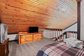 Een slaapkamer met een houten plafond, een televisie op een dressoir, een ventilator, een bed met een ruitdessin en een trap.