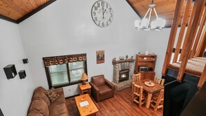 A cozy living room with a stone fireplace, wooden furniture, a clock, and a window with blinds.