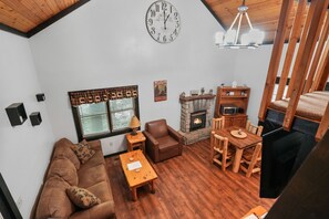 Un salon chaleureux avec une cheminée en pierre, des meubles en bois, une horloge et une fenêtre avec des stores.
