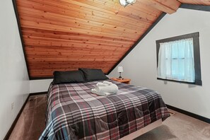 Une chambre avec un plafond en bois, une couvre-lit à carreaux, un coussin noir, une serviette blanche et une fenêtre ornée de rideaux légers.