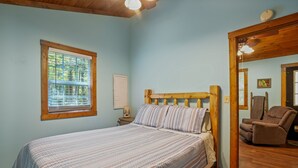 Ein Schlafzimmer mit einem Bett, einem Fenster mit Jalousien, einem Sessel und einer Holzbalkendecke.