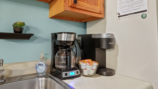 Eine Küche mit Spüle aus Edelstahl, einer Kaffeemaschine, einem Wasserspender und einer Schüssel aus Glas mit orangen Gegenständen.