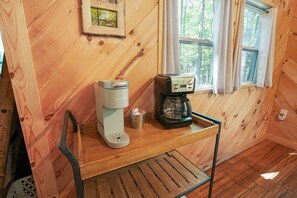 L’intérieur d’un chalet en bois, avec une machine à café posée sur une table en bois, un tableau encadré accroché au mur et une fenêtre avec des rideaux.