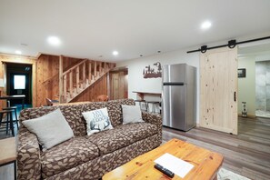 Ein modernes Wohnzimmer mit einem gemusterten Sofa, einem Holztisch, einer Küchenzeile inklusive Kühlschrank und einer Treppe.